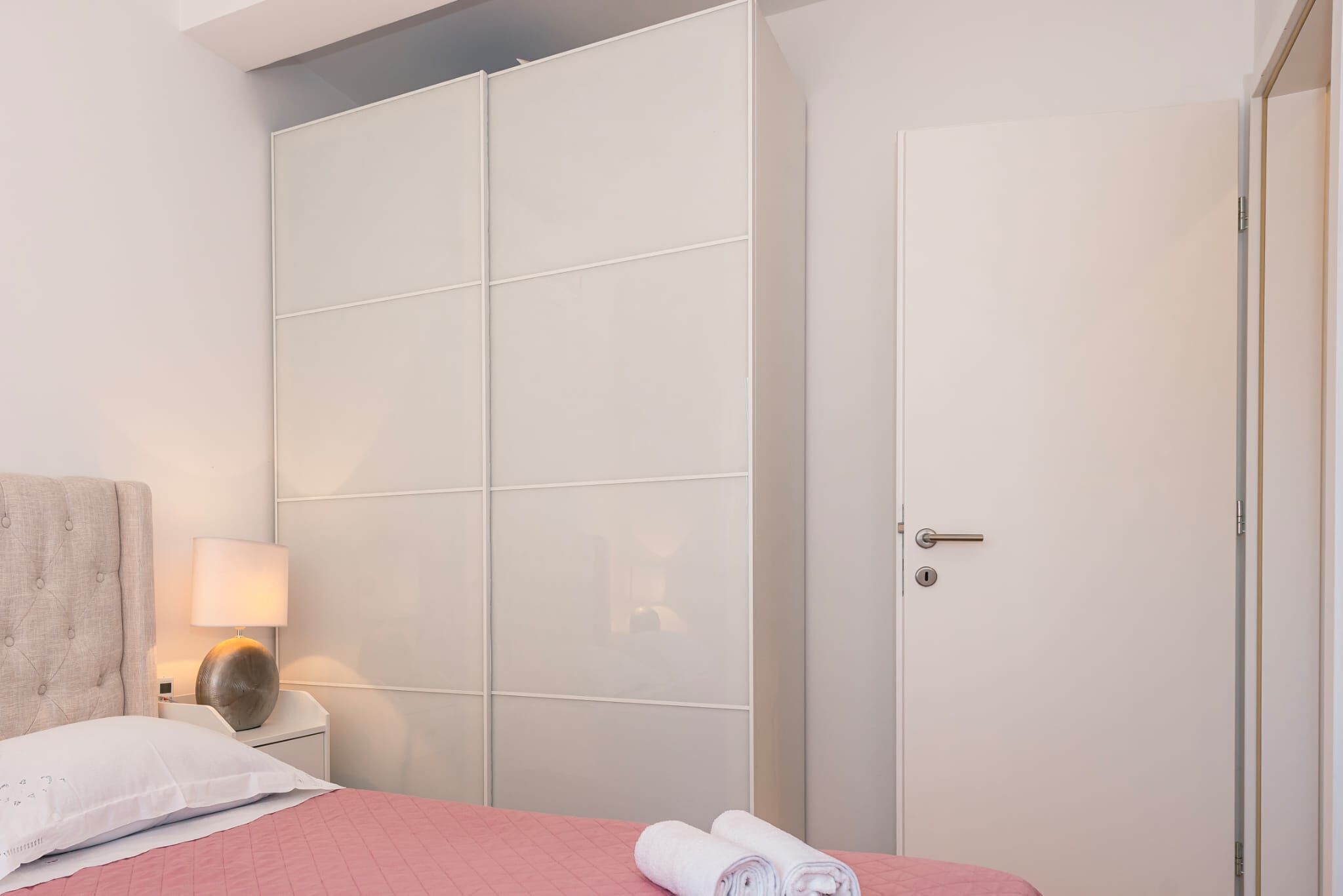A bedroom with a bed, a nightstand, a lamp, a wardrobe, and a door.