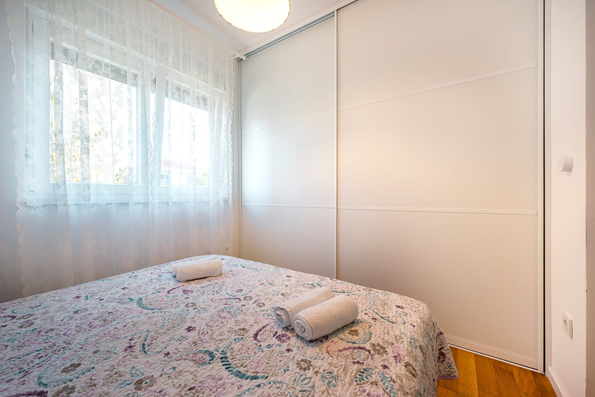 A bedroom with a bed, a window with sheer curtains, and a white wardrobe.