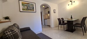 Egy modern nappali, amelyben van egy kanapé, egy étkezőasztal székekkel, a háttérben egy konyha, és a falon egy kép, amely keretben van.