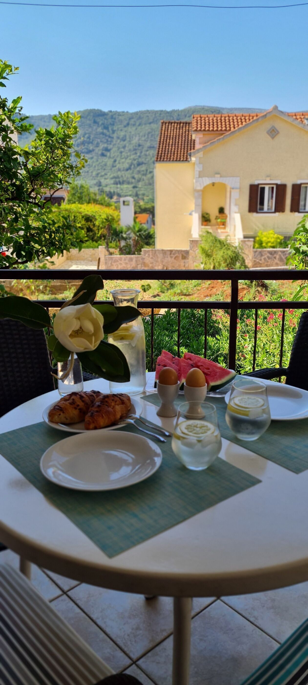 Une table dressée avec des croissants, une assiette de pastèque et des verres d’eau, le tout sur un balcon d’où l’on peut admirer une maison et de la verdure.