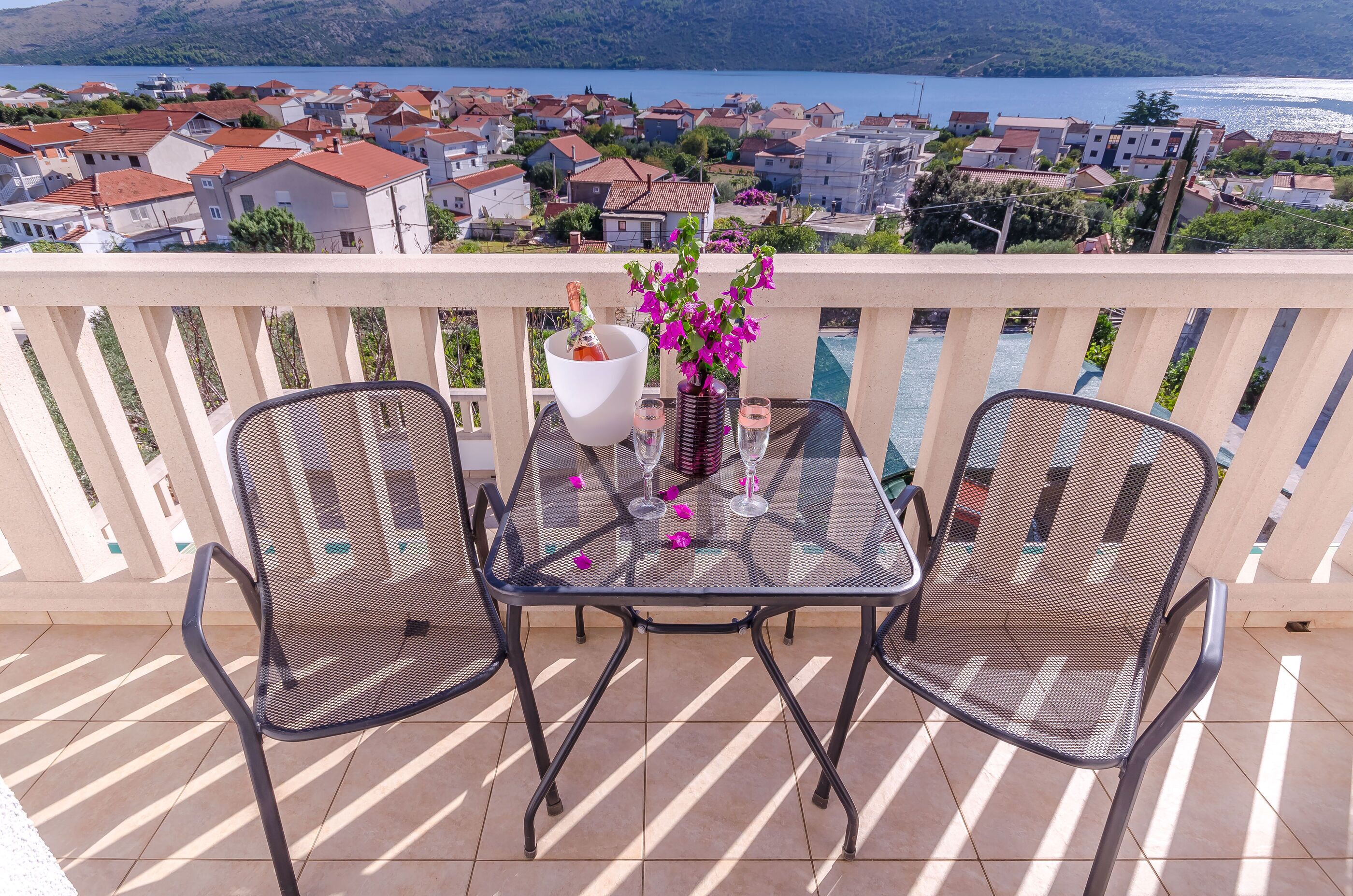 A balcony with a table and two chairs, overlooking a coastal town with houses and a body of water.