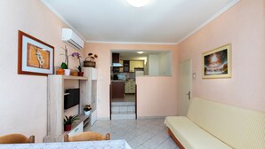 A living room with a sofa, a dining table with a lace tablecloth, a TV, and a kitchen in the background. - House Nenad (Lopar)