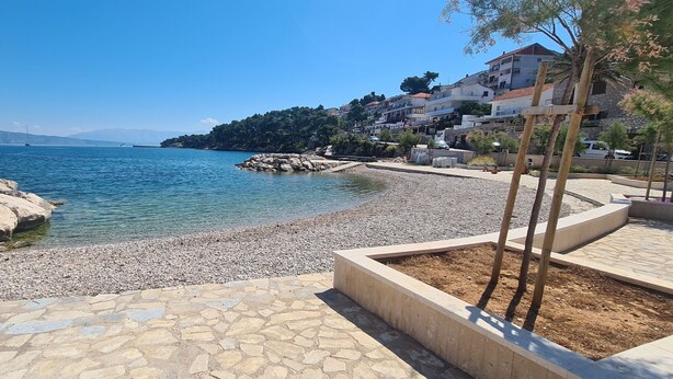 A coastal area with a pebble beach, clear blue water, and buildings in the background. - Apartment Bosiljak (Jelsa)