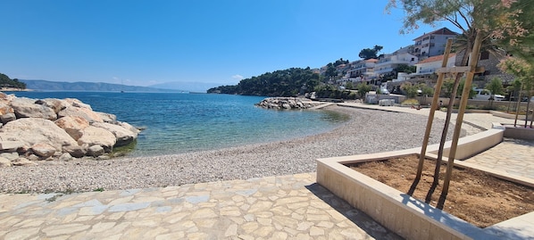 A coastal area with a pebble beach, clear blue water, and buildings in the background. - Apartment Bosiljak (Jelsa)