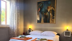 A bedroom with a wooden herringbone floor, a bed with white bedding, two bedside lamps, a painting on the wall, and a window with curtains. - Apartment Bosiljak (Jelsa)