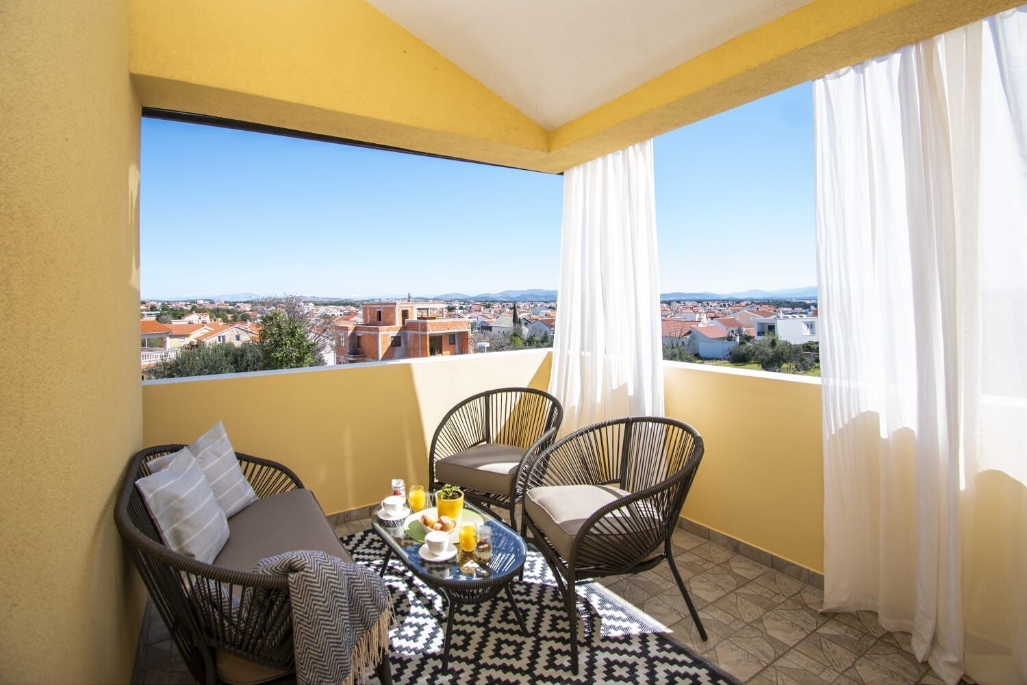 Varanda com mesa posta para o café da manhã, duas cadeiras de vime e vista para uma cidade.