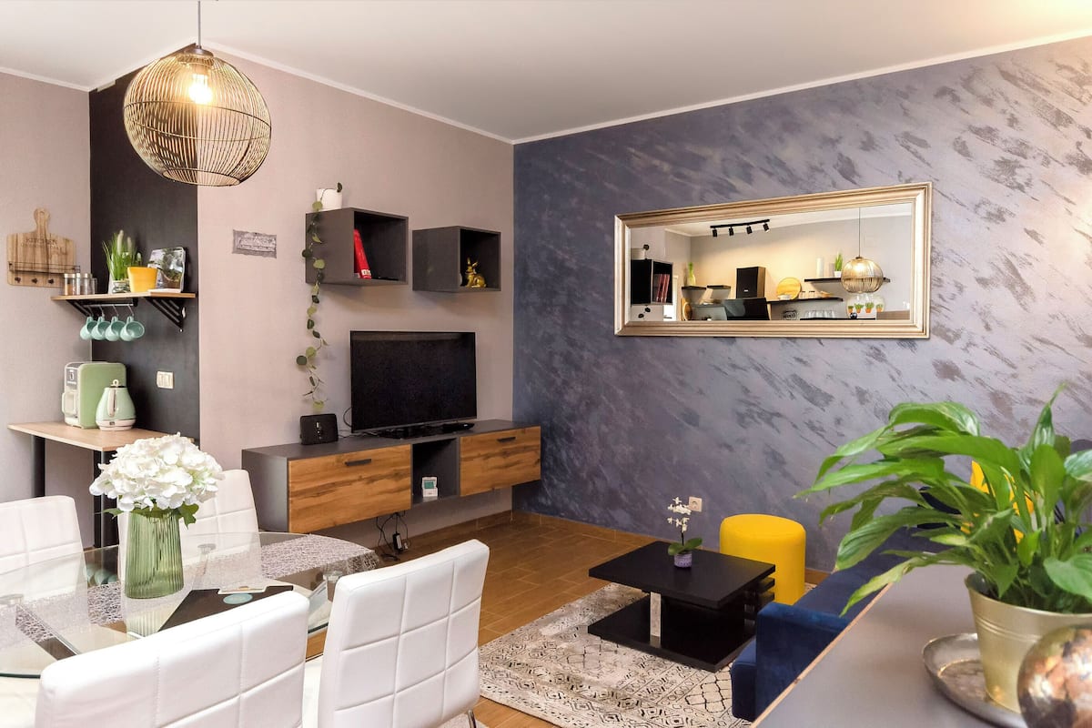 Ein modernes Wohnzimmer mit Fernseher, Holzschränken, einem Spiegel und einem Essbereich mit Glastisch und weißen Stühlen.