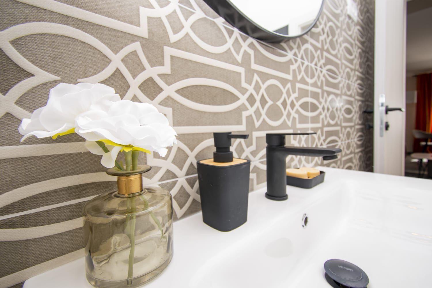 Un baño con un lavamanos blanco, un espejo, una botella de perfume y un dispensador de jabón.