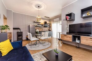 Ein modernes Wohnzimmer mit einer Couch, einem Couchtisch und einem Fernseher. Im Hintergrund befindet sich eine Essecke mit Stühlen und einem Tisch sowie eine Küche.