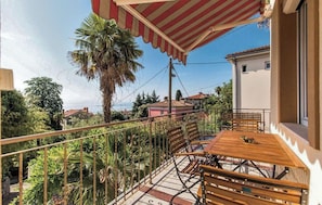 A balcony with a wooden table and chairs, a striped awning, and a view of the sea and surrounding houses. - Apartments Villa Viktorija (Opatija)