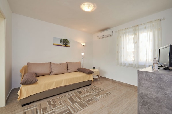 A modern bedroom with a sofa bed, a small bedside table with a lamp, a TV, and a window with sheer curtains. - House Dvor (Seget)