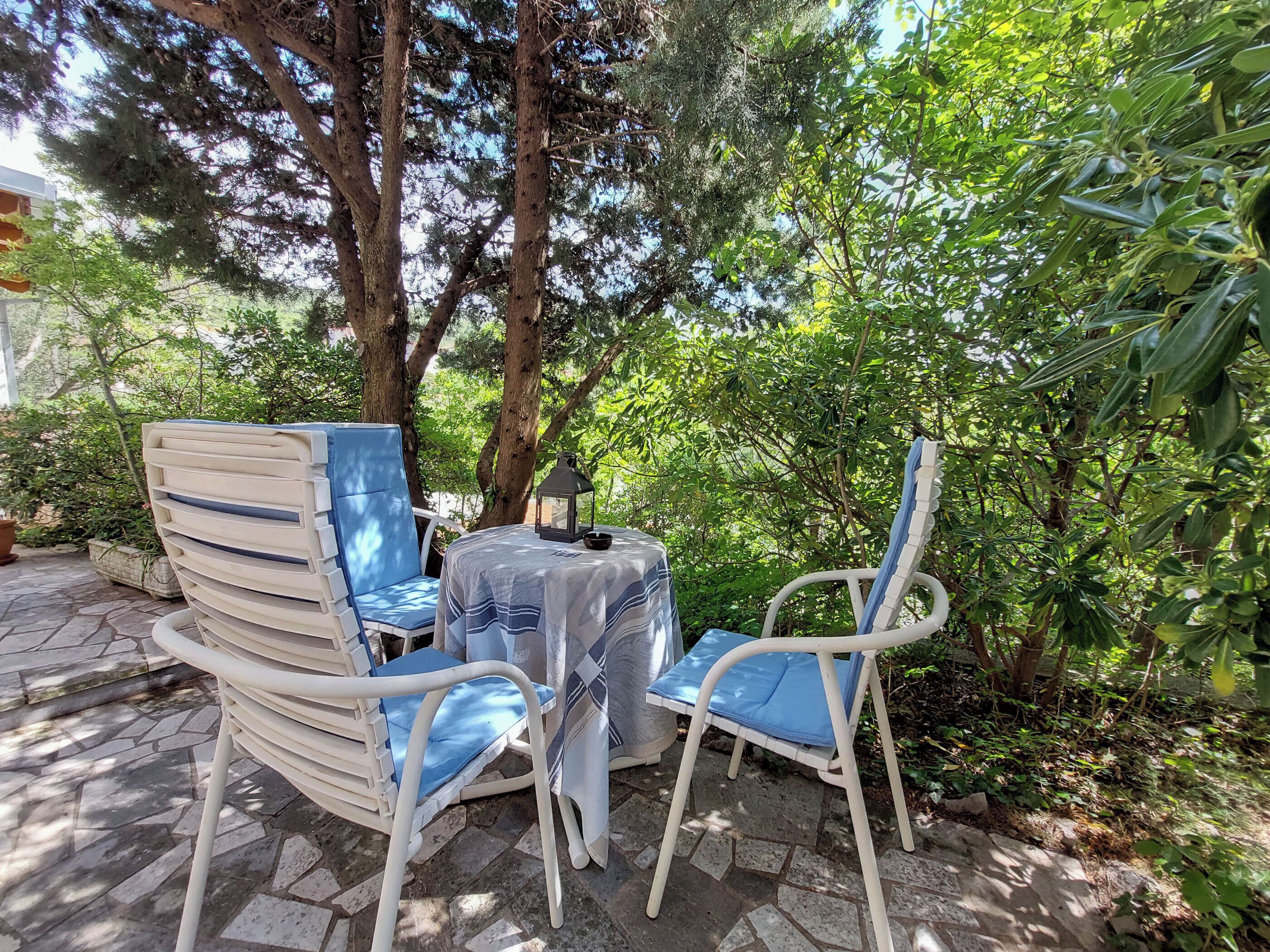 An outdoor seating area with two white chairs and a table, surrounded by greenery and trees.