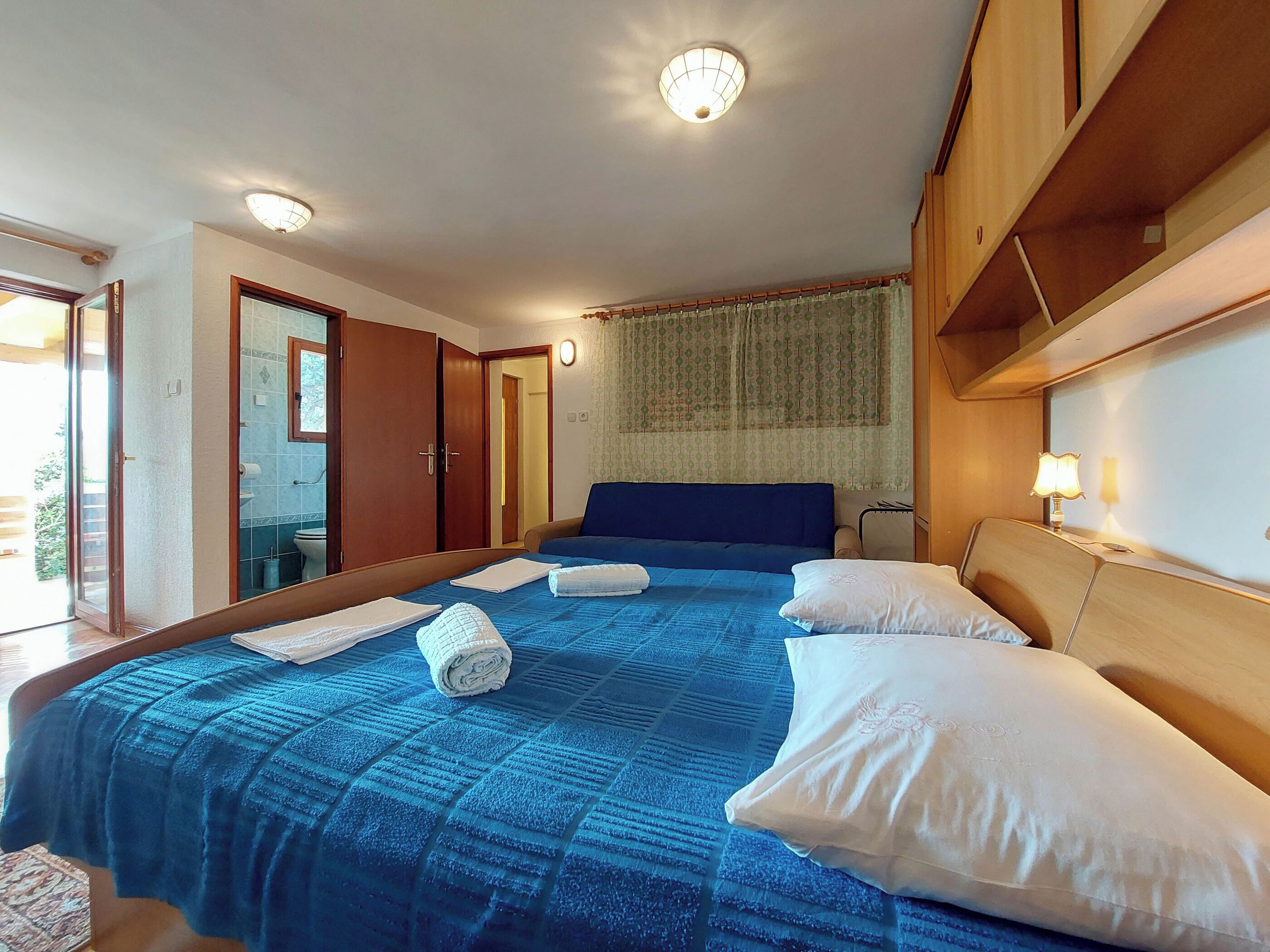 A hotel room with a large bed, a wooden headboard, a nightstand with a lamp, and a bathroom visible through an open door.