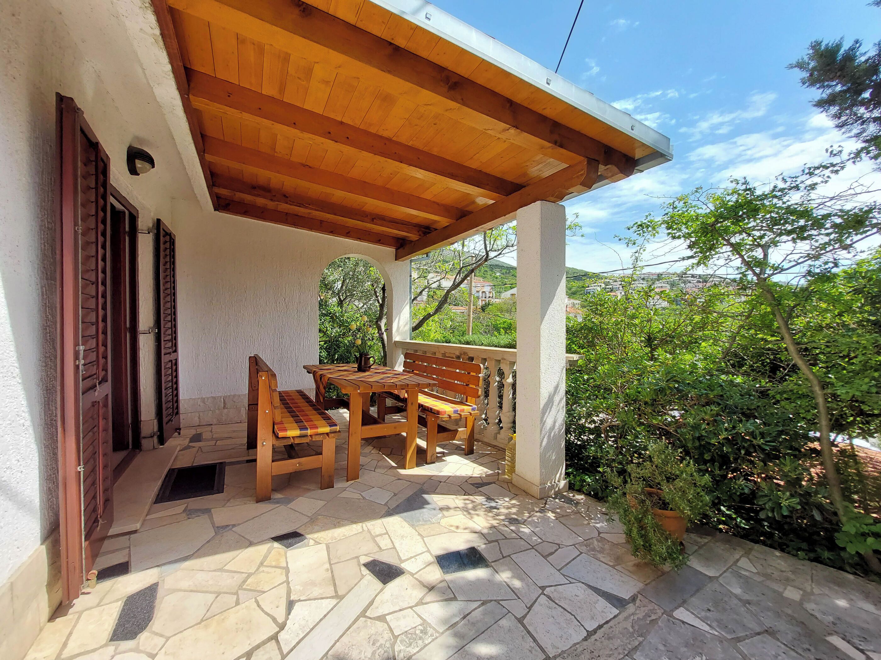 A covered patio with a wooden table and chairs, stone flooring, and a view of greenery and buildings in the background.
