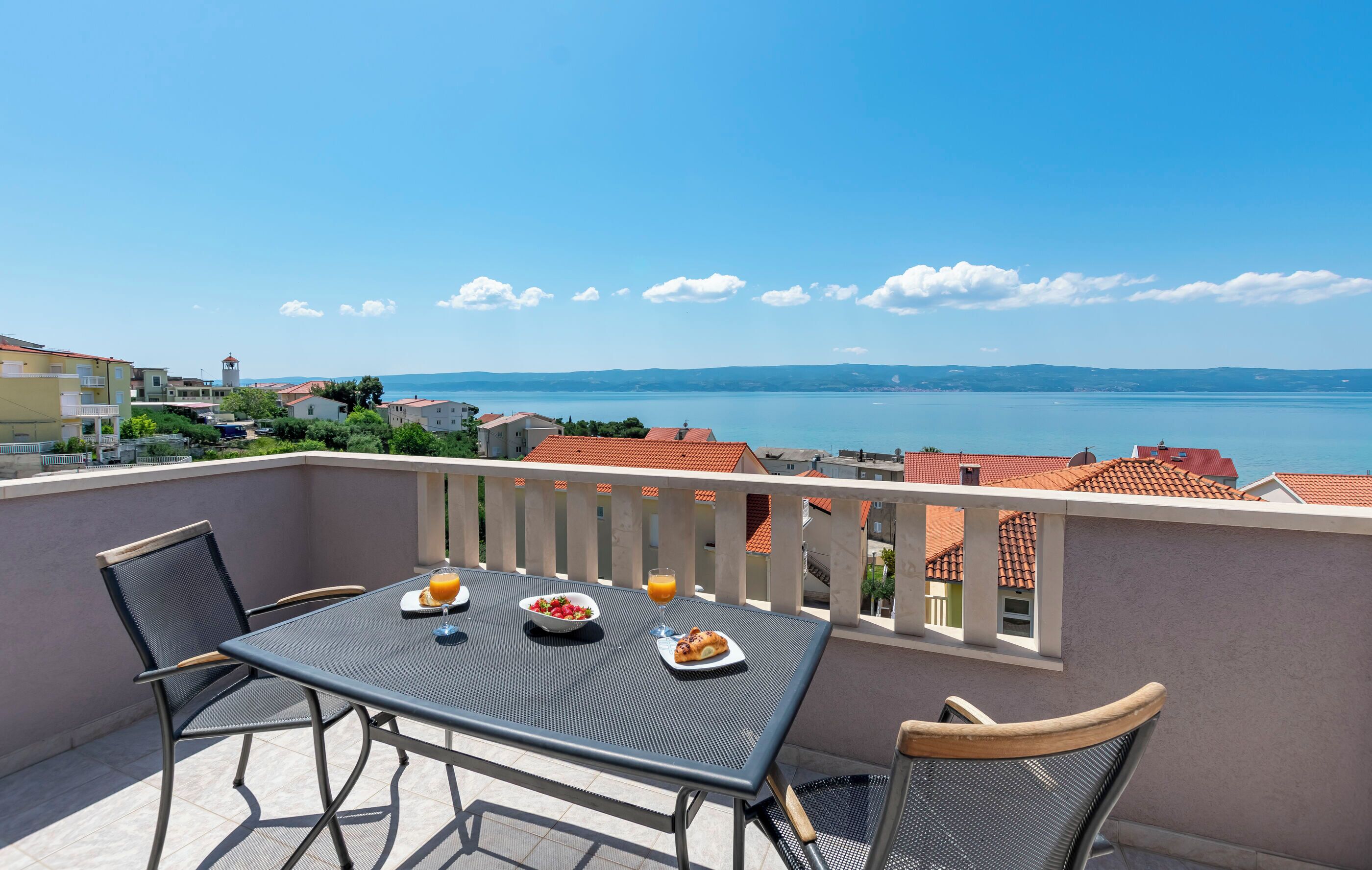 A balcony with a table set for breakfast, overlooking a coastal town and sea.