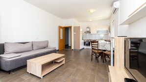Un apartamento moderno con un sofá gris, una mesa de centro de madera, una zona de comedor con mesa y sillas, y una cocina con armarios blancos y una estufa.