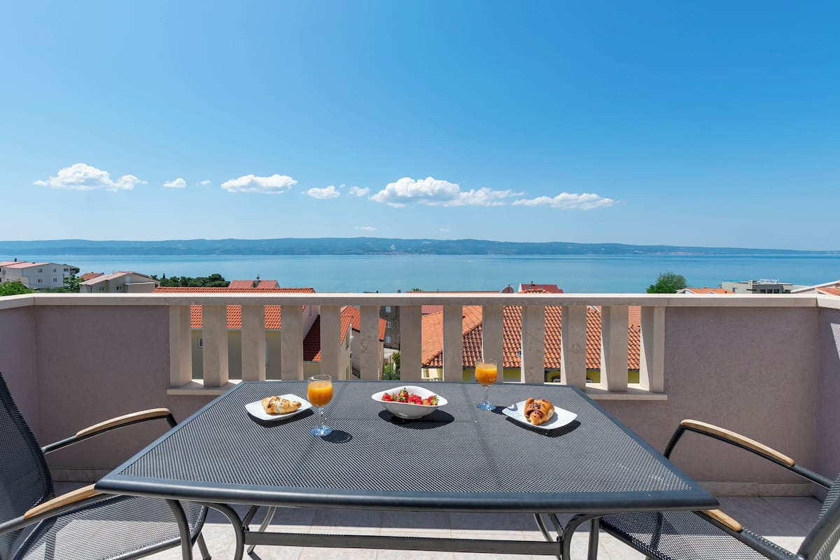 En balkong med ett bord dukat för frukost, med utsikt över ett vackert landskap med havet och avlägsna berg.