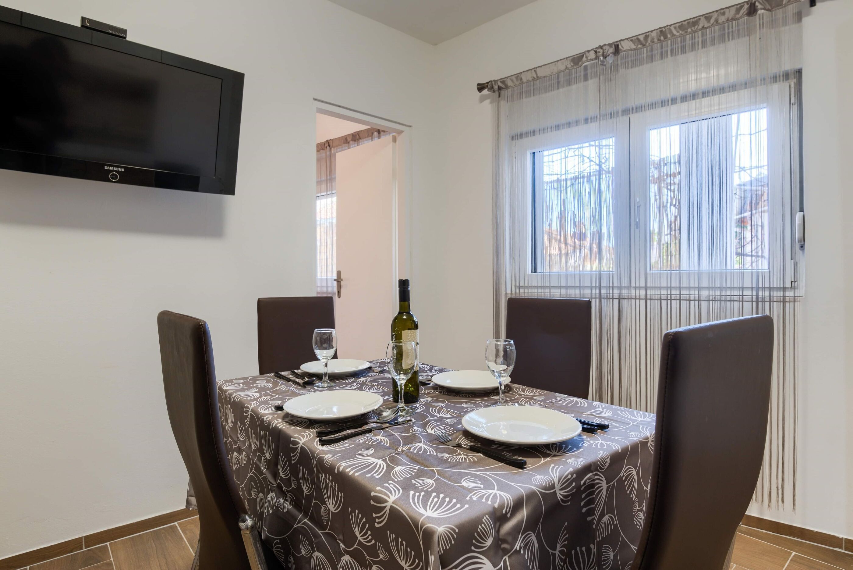 Un comedor con una mesa preparada para cuatro, una botella de vino y un televisor de pantalla plana montado en la pared.