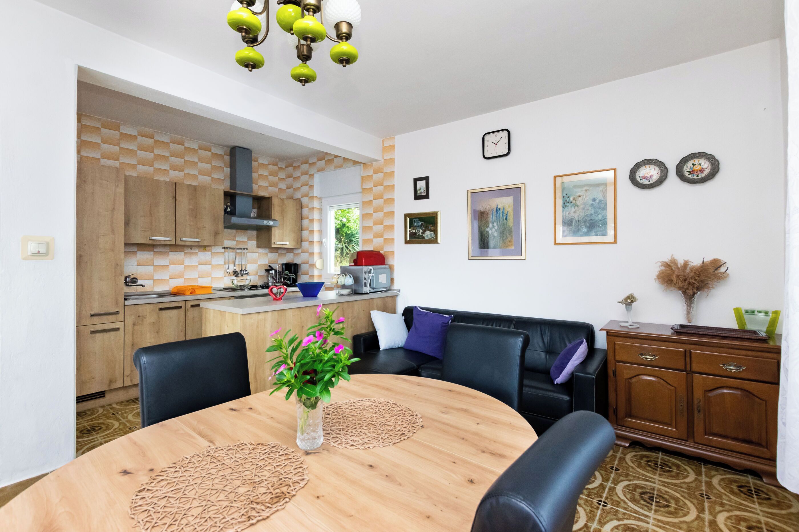 A modern living room with a round wooden table, black leather sofa, and a dining area with a checkered tile backsplash.