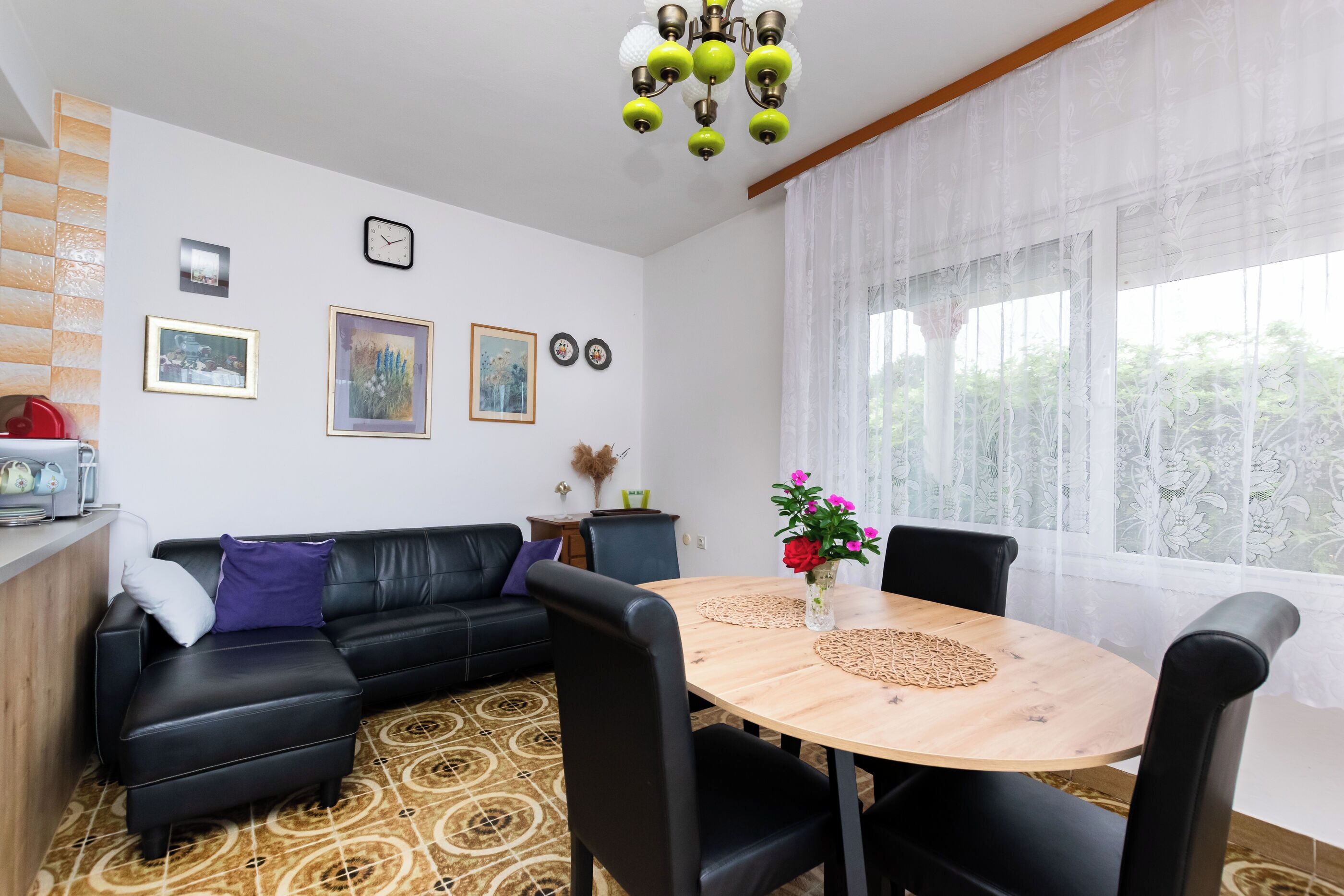 Un salon moderne avec un canapé en cuir noir, une table à manger en bois et quatre chaises, un lustre et une grande fenêtre ornée de rideaux légers.