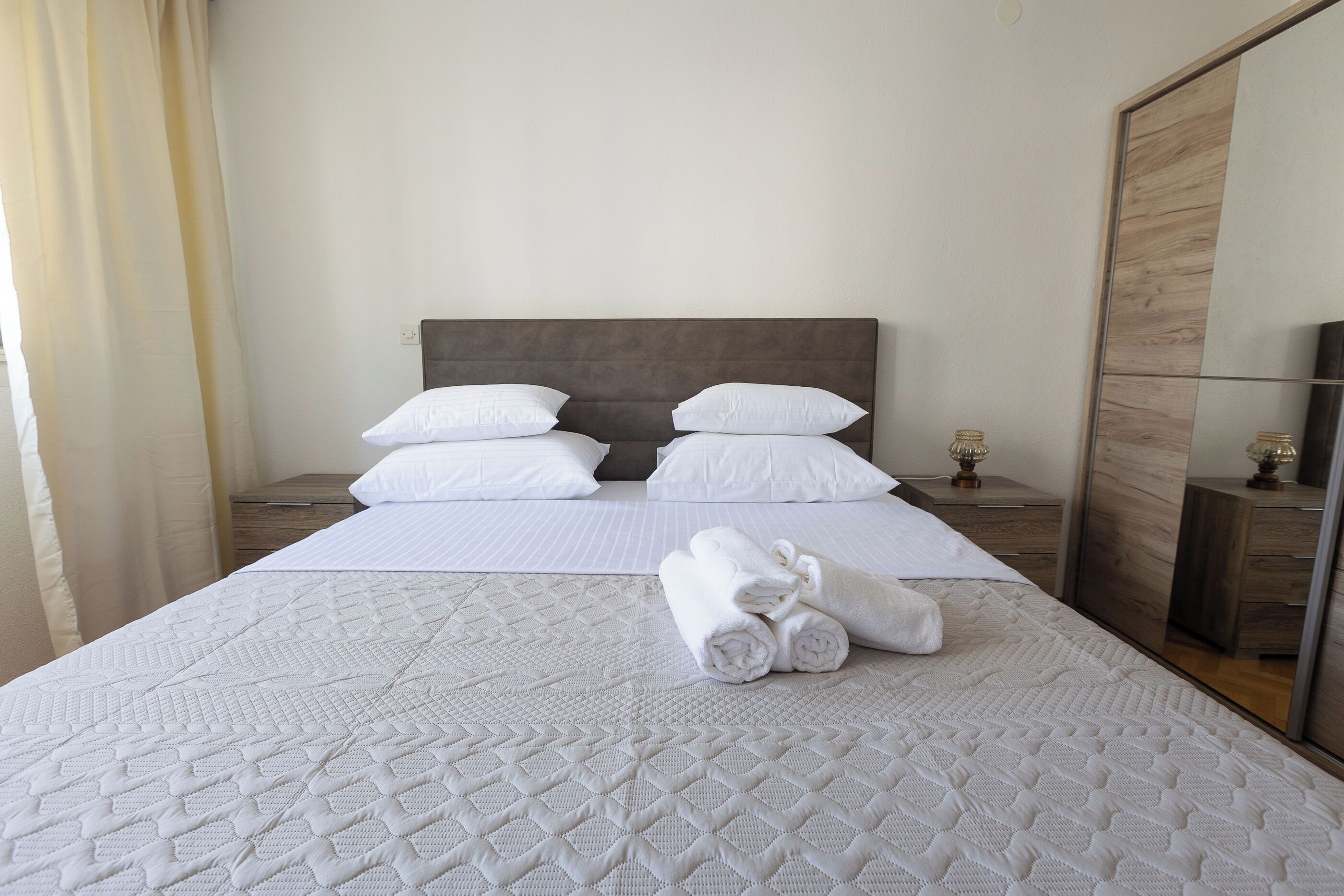 Een opgemaakt bed met witte lakens en gerolde handdoeken, een houten hoofdbord en een spiegelkast.