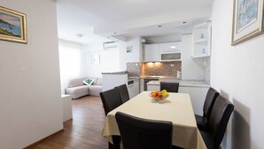 Et moderne køkken med hvide skabe, et spisebord med en skål frugt og et opholdsområde med en sofa og et billede.