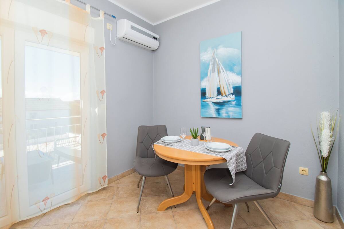 A dining area with a round table, two grey chairs, a vase with white flowers, and a painting of a sailboat on the wall.