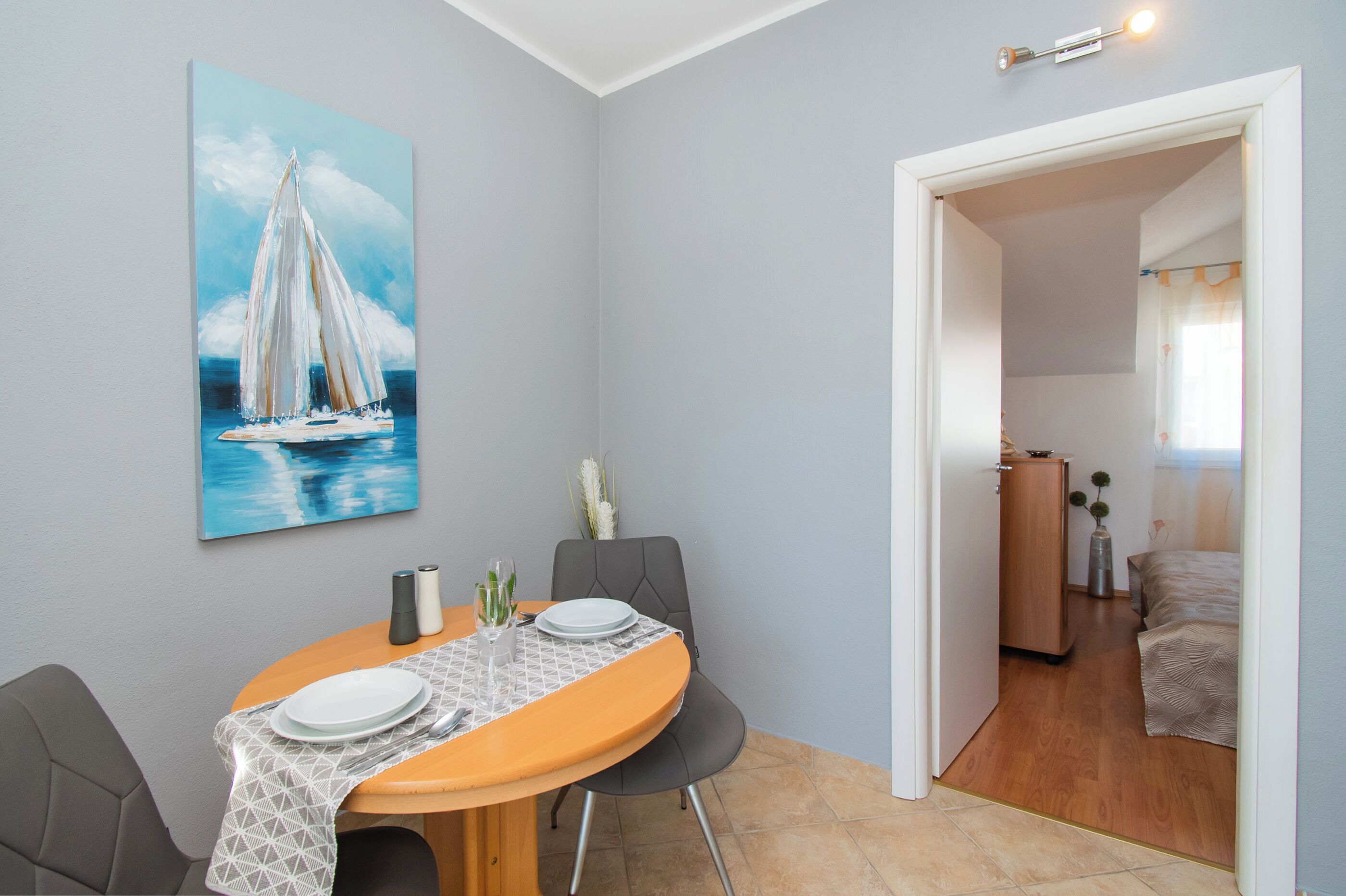 Un comedor con una mesa dispuesta para dos, un cuadro de un velero en la pared y vistas a un dormitorio a través de una puerta abierta.