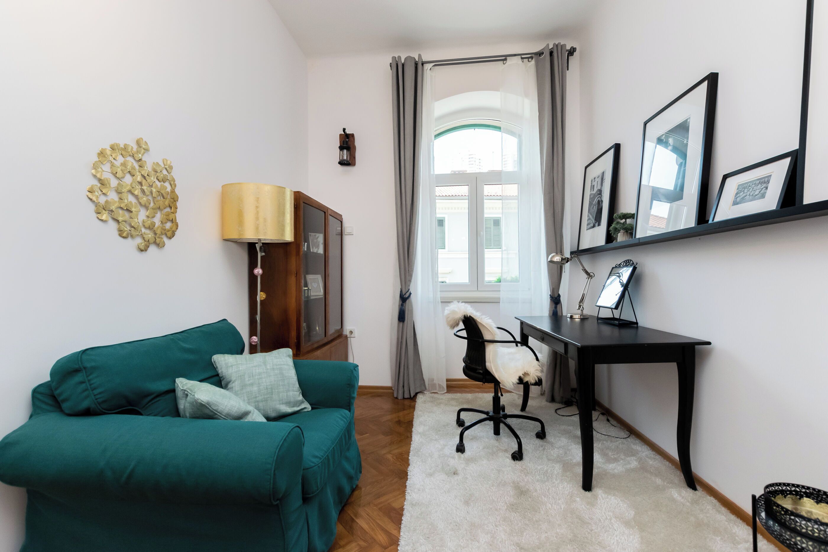 A living room with a teal sofa, a white rug, a wooden cabinet, a desk with a lamp, and a window with curtains.
