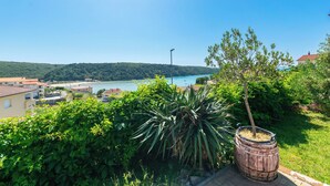 A coastal view with a wooden barrel planter, lush greenery, and a distant body of water. - Apartments Zeljko (Rab)