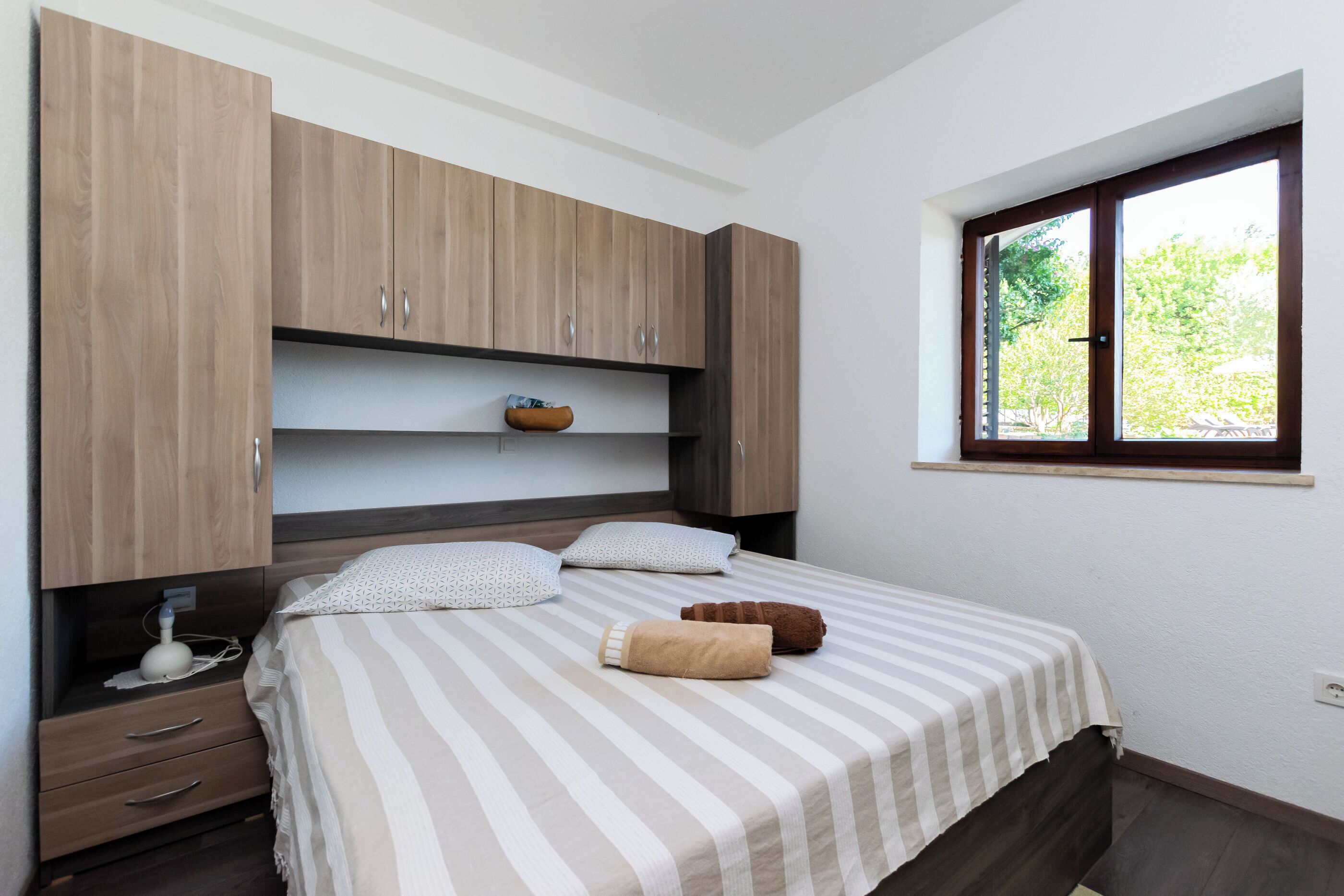 Un dormitorio con cama, armarios de madera, una mesita de noche y una ventana con vista a follaje.