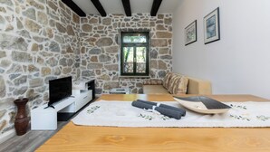 A cozy room with a stone wall, a wooden table, a sofa, a TV, and a window with a view of greenery. - Apartments Zeljko (Rab)