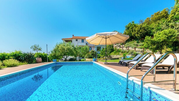 A swimming pool with lounge chairs and an umbrella, surrounded by greenery and a building in the background. - Apartments Zeljko (Rab)