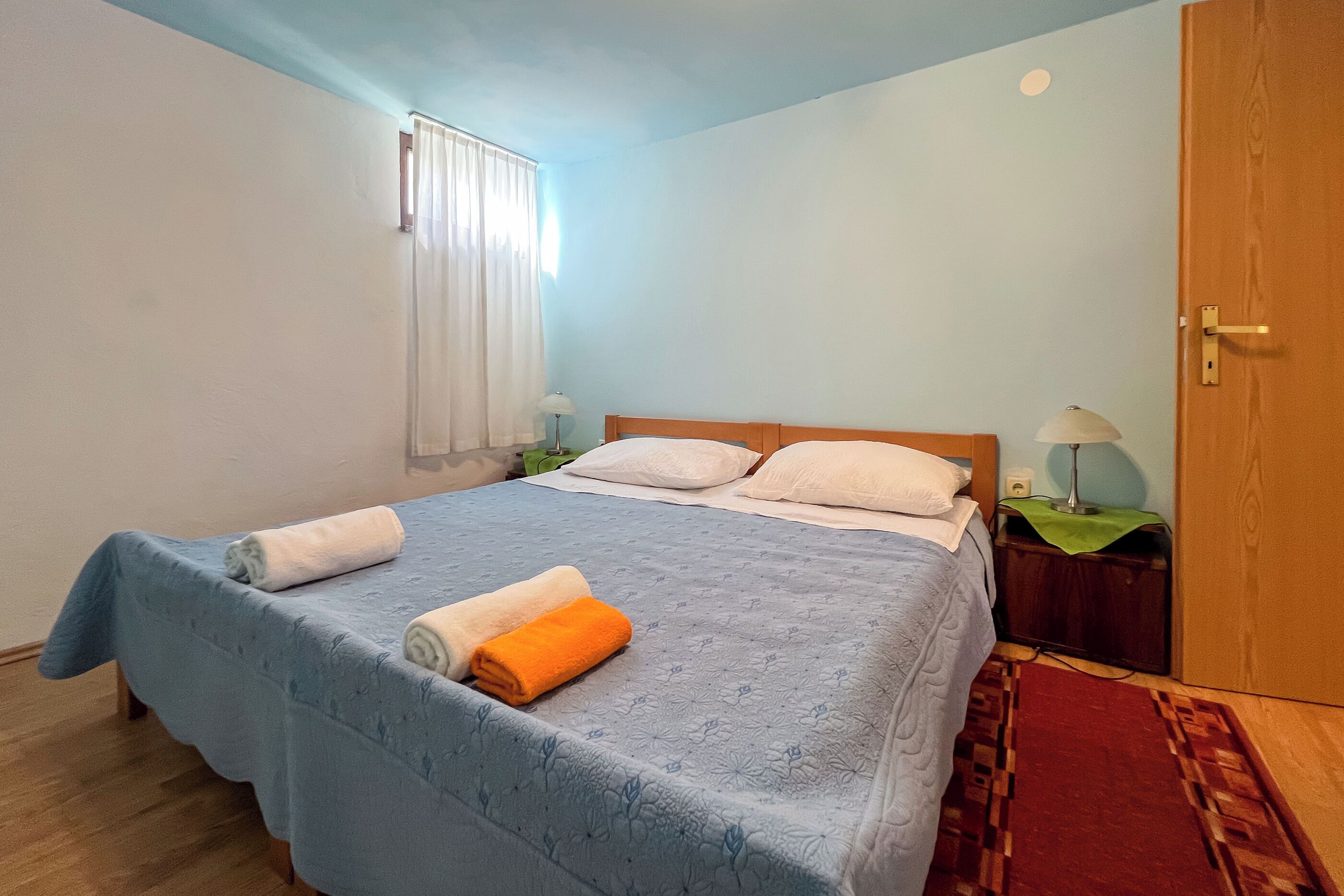 A neatly made bed with a blue bedspread, two pillows, and rolled towels.