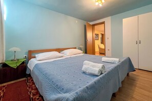 A bedroom with a large bed, two bedside tables, a nightstand, a wooden wardrobe, and a door leading to another room. - Apartments Franjo (Rab)