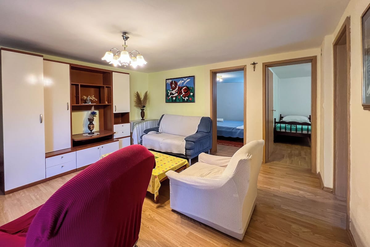 Un salon comprenant un canapé, des fauteuils, une table basse et une bibliothèque. Un tableau est accroché au mur et une porte donne accès à une autre pièce.