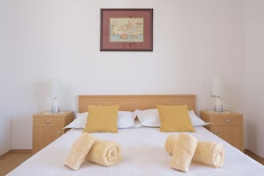 Een net opgemaakt bed met witte en mosterdgele kussens, twee opgerolde handdoeken en een ingelijste foto erboven.