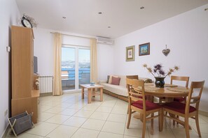 Ein Wohnzimmer mit Sofa, Esstisch mit Stühlen, Holzschrank und Blick durch ein großes Fenster.