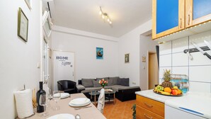 A modern kitchen with white cabinets, a countertop with fruits, and a dining area with a table set for two. - Apartment Vlatka Grof (Crikvenica)