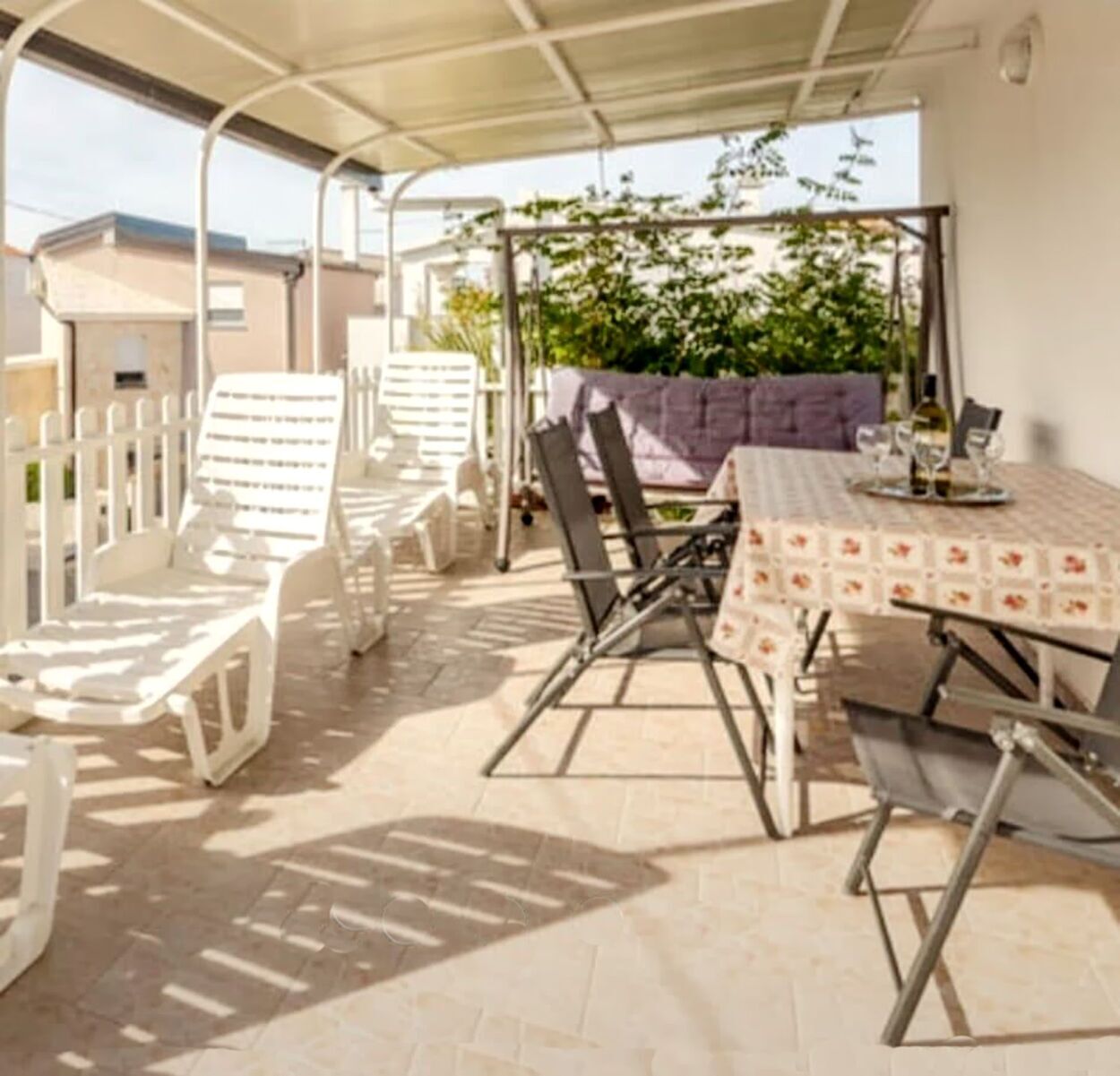 Een terras buiten met een tafel, stoelen, een pergola en uitzicht op gebouwen en groen.