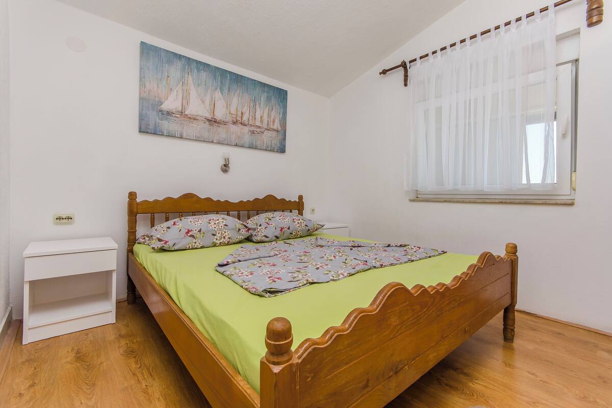 A neatly made bed with a floral bedspread and pillows, a white nightstand, a painting of sailboats on the wall, and a window with sheer curtains.