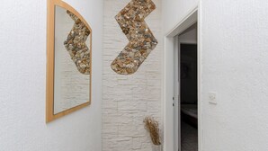 Ein Korridor mit einer weißen, strukturierten Wand, einem spiegelnden Rahmen aus Holz und einem dekorativen Wandelement aus Steinen.