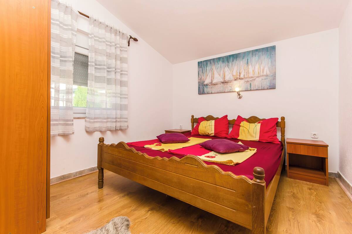 A bedroom with a wooden bed, a nightstand, a window with sheer curtains, and a painting on the wall.