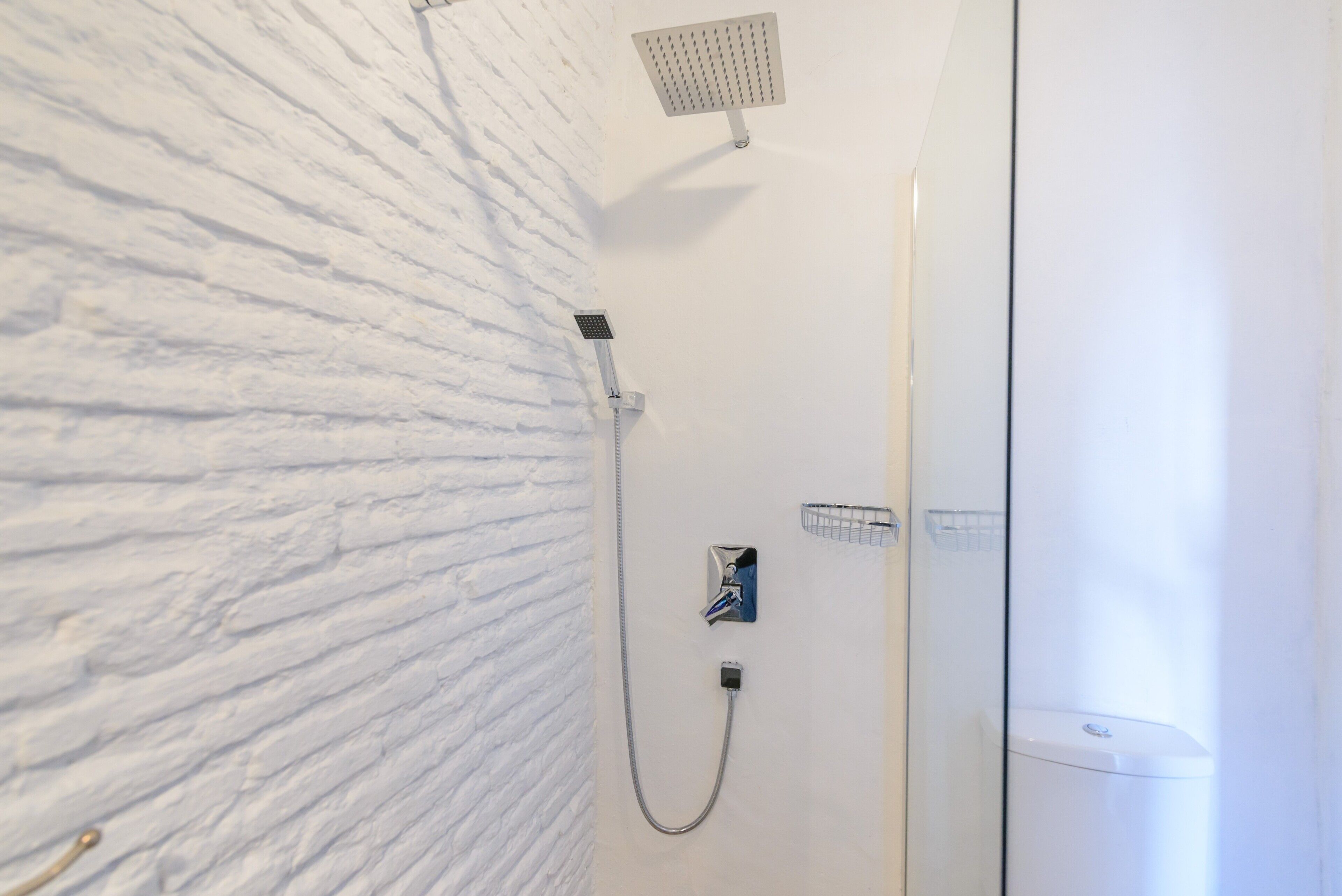 A shower with a white textured wall, a handheld showerhead, a wall-mounted faucet, and a toilet.