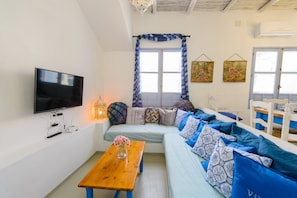 A living room with a flat-screen TV, a wooden table with a vase, a sofa with patterned cushions, and a window with curtains. - Casa de la Tita Vitoria: Rural House with Mountain Views, Wi-Fi and Air Conditioning (La Puebla de los Infantes)