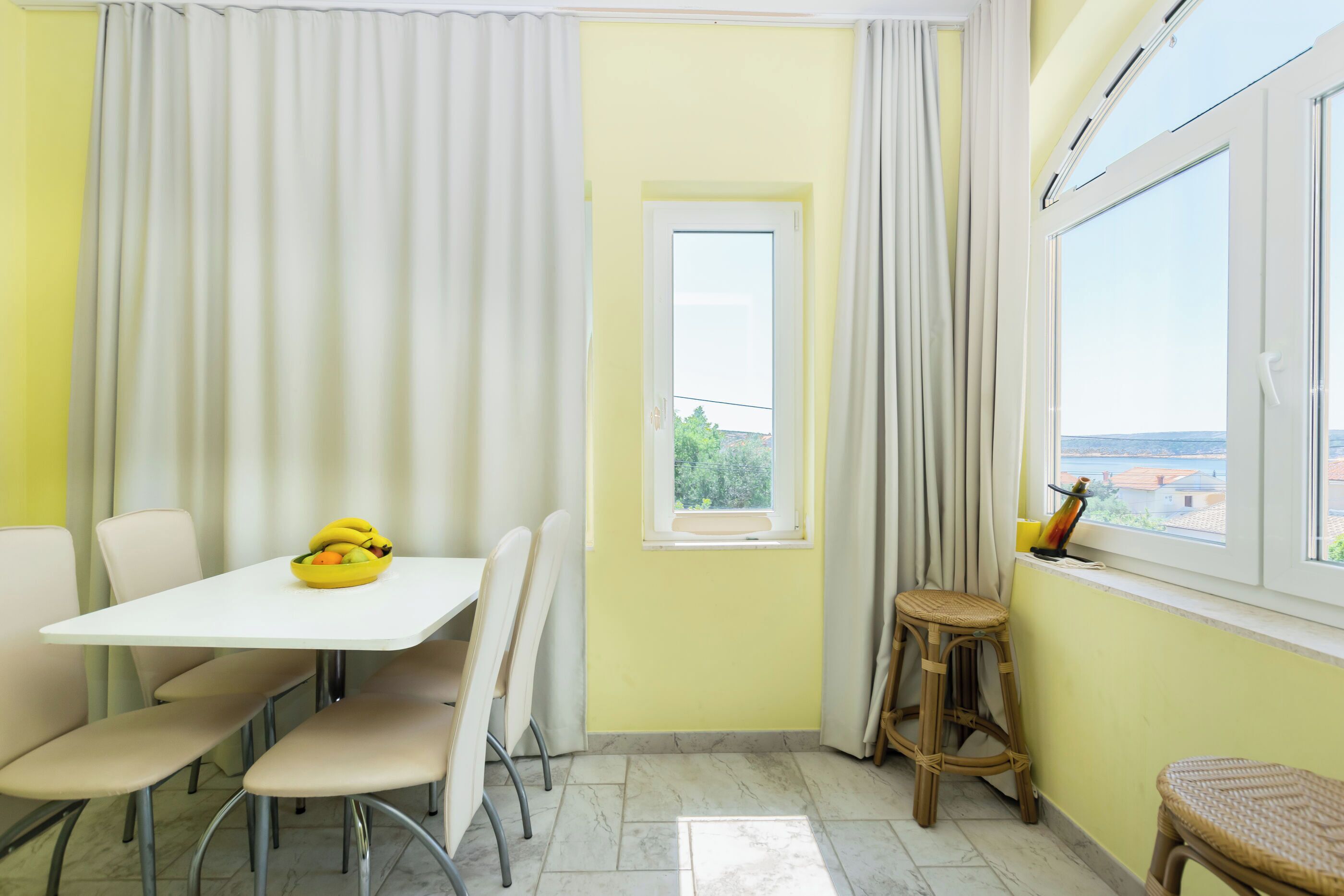 A corner room with a white table, four chairs, a small window, and a view of the outside.