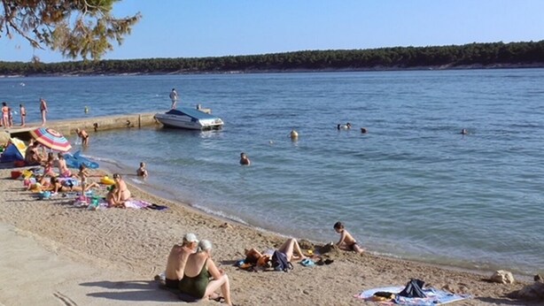 A beach with people swimming and sunbathing, a boat docked near the shore, and a clear sky. - Apartments Rada i Stanislav (Rab)