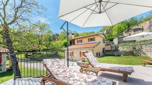 Eine Terrasse mit zwei Liegestühlen und einem großen Sonnenschirm, eine Steinmauer und im Hintergrund ein Haus mit einem Ziegeldach.