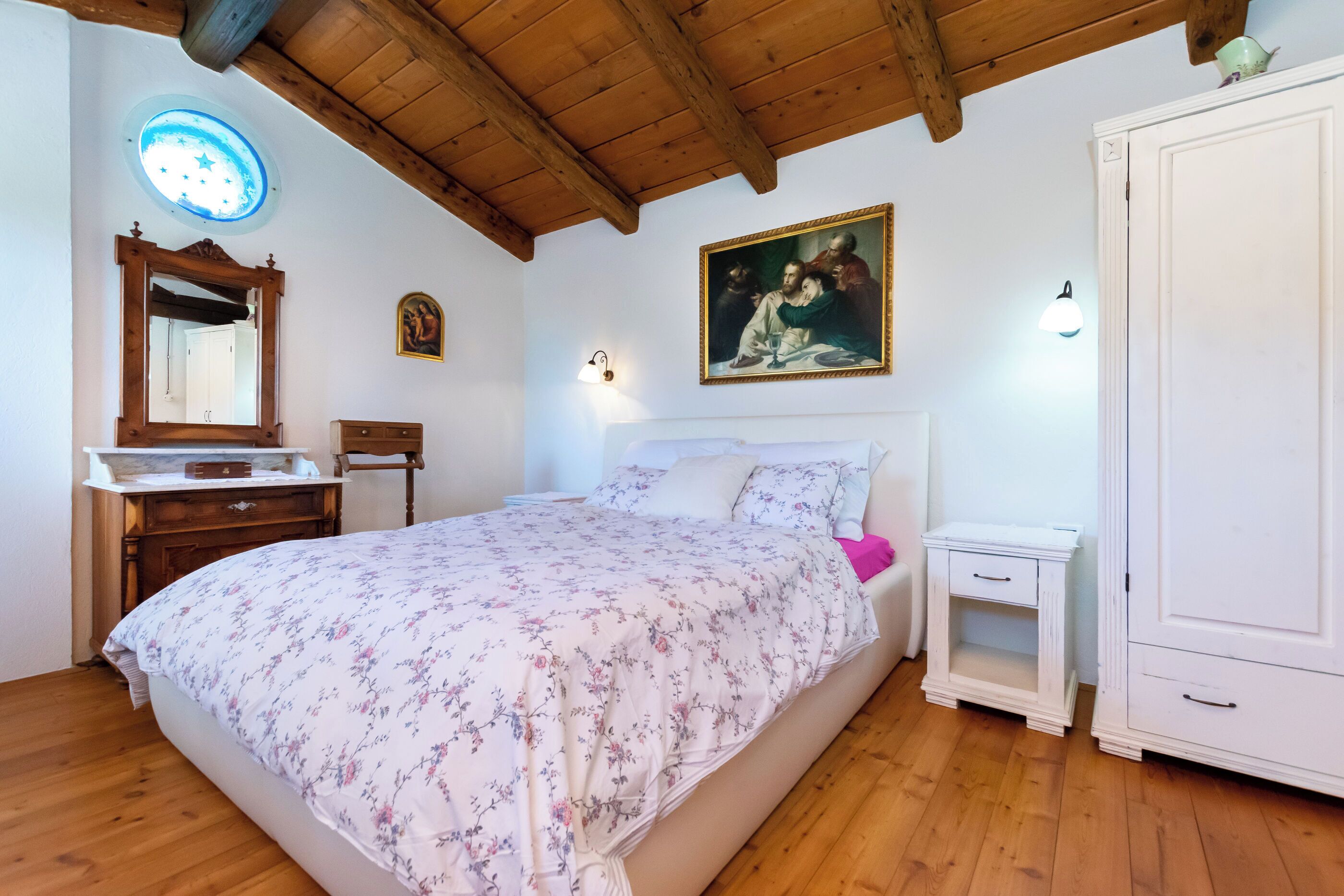 A bedroom with a wooden ceiling, a bed with floral bedding, a nightstand, a dresser, a mirror, and a painting on the wall.