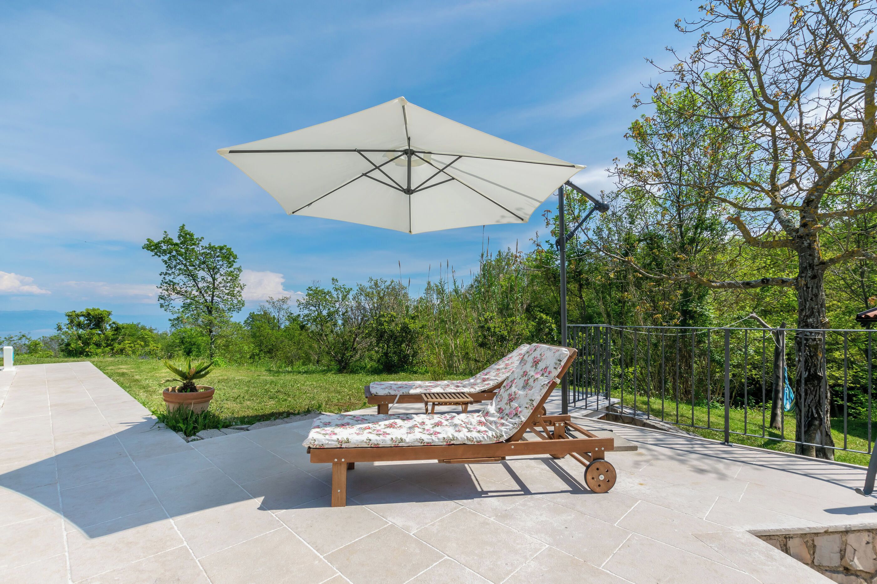 Two wooden lounge chairs with floral upholstery and a large umbrella, set on a paved patio with a view of greenery and trees.